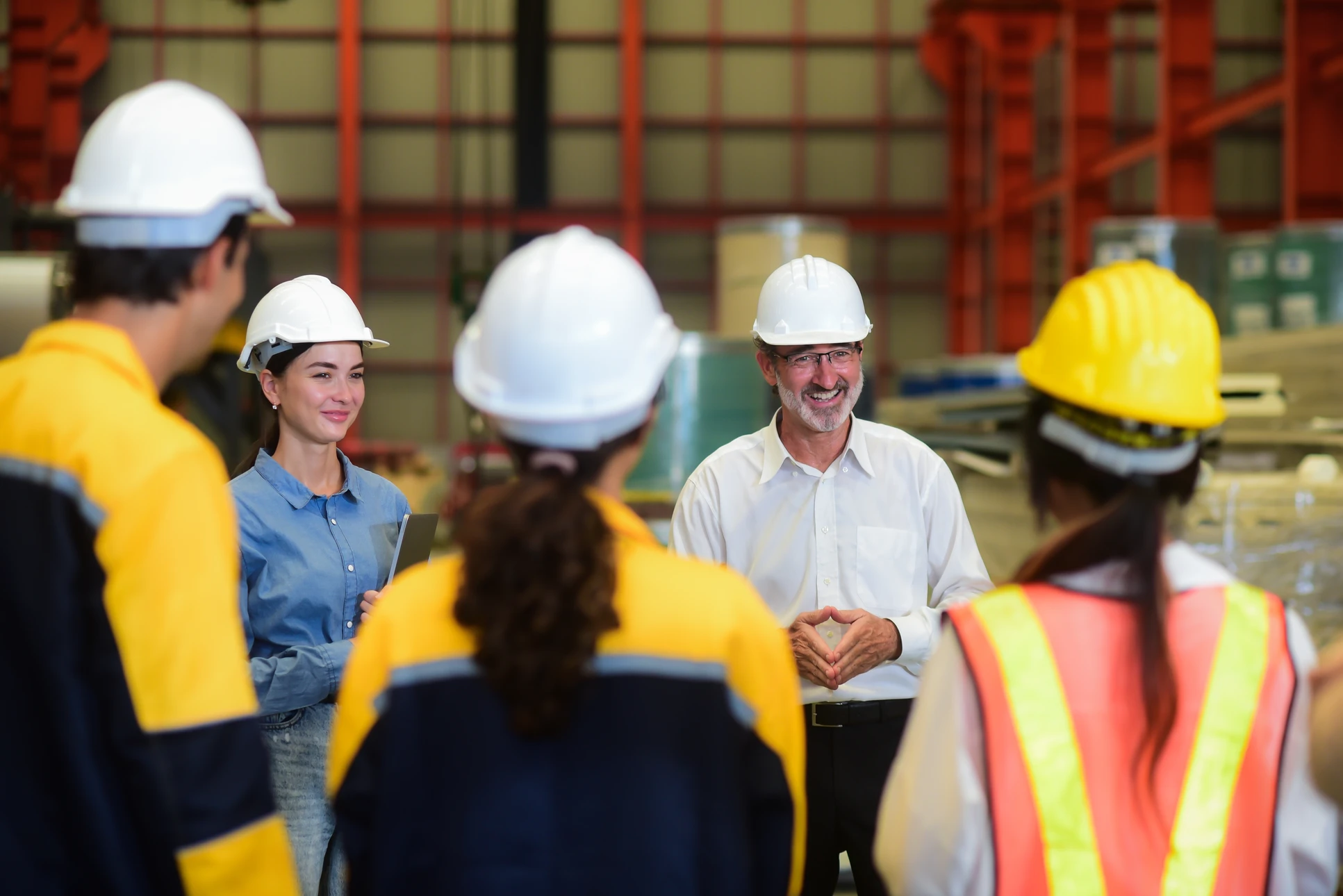 Réunion d'équipe dans un entrepôt industriel avec managers et ouvriers portant casques de sécurité blancs et jaunes, gilets haute visibilité